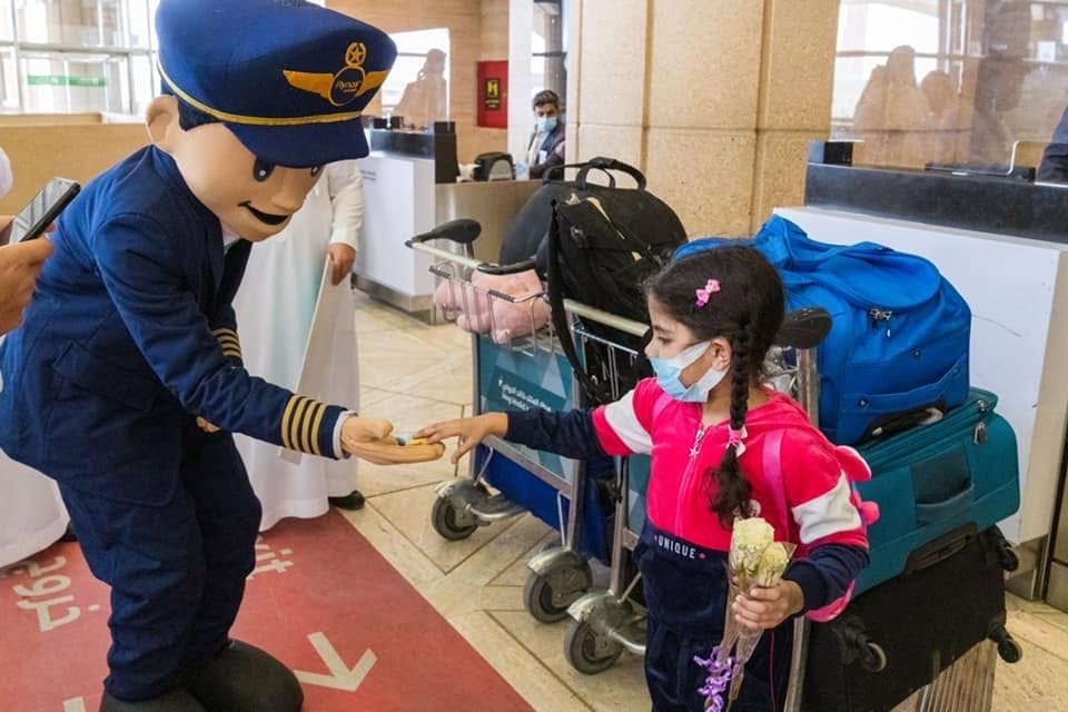 Captain Firnas mascot handing a small gift to a young traveler at the airport